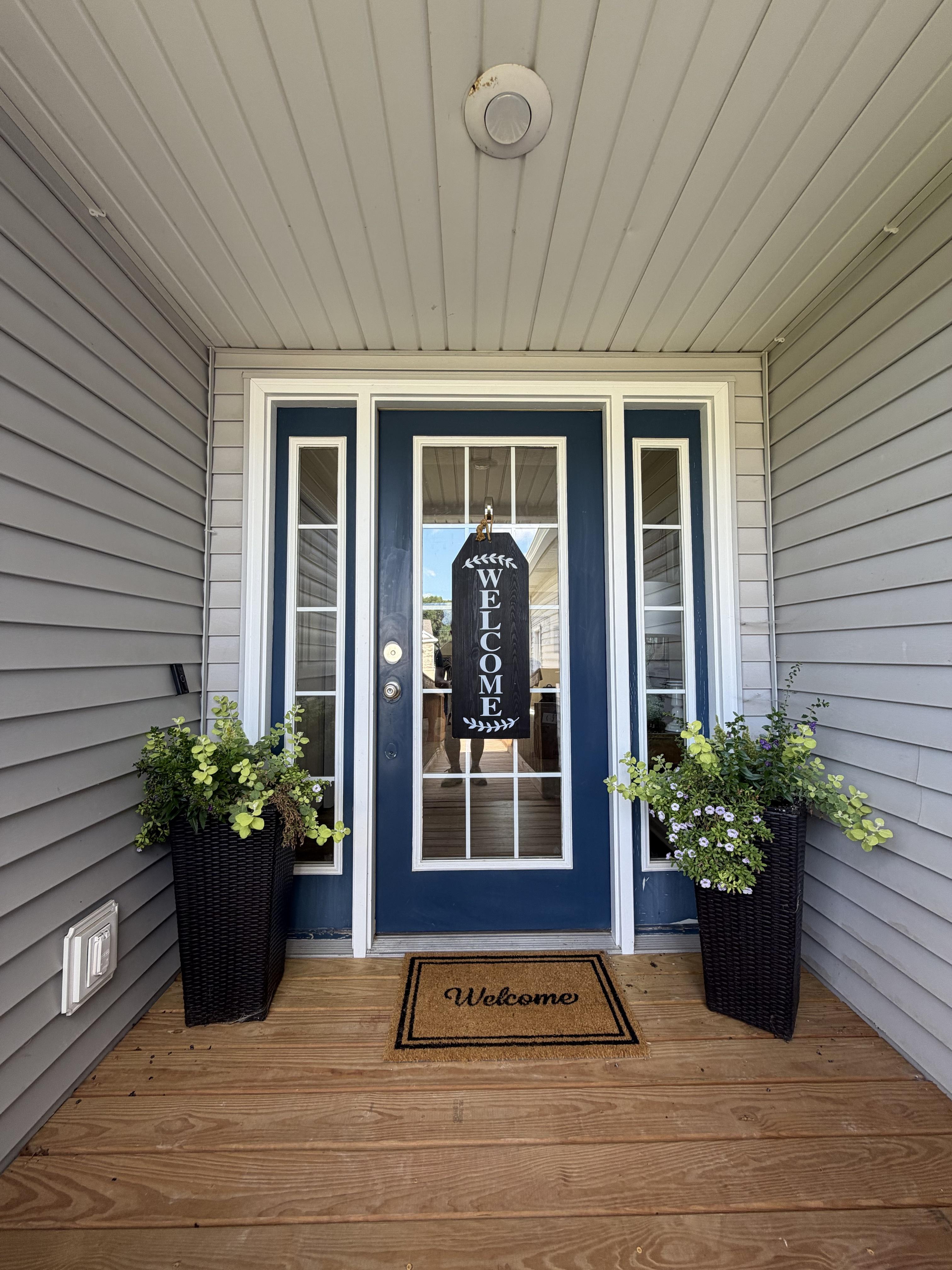 Covered front porch with a navy entry door, a Welcome sign, and potted flowers